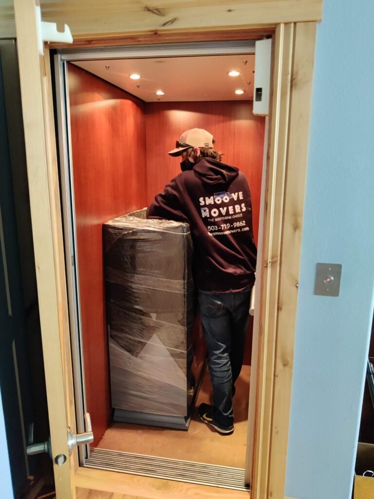 Home Elevator - The Smoove Movers - The Smoove Movers Mover in a Smoove Movers hoodie inside a small elevator, guiding a tall, wrapped appliance.