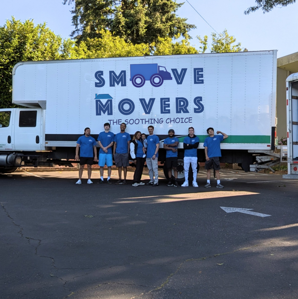 Team Photo - The Smoove Movers Group of movers in blue shirts standing in front of a large white moving truck with'MOVERS' lettering and a blue truck logo, outdoors on a sunny day
